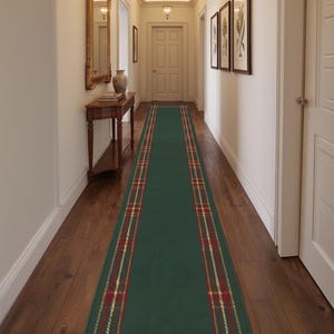 May include: A long rectangular runner rug featuring a solid dark green center panel framed by a decorative border with a red, gold, and white plaid pattern. The rug is positioned lengthwise along the center of a wooden floor in a hallway.