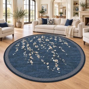May include: A large circular area rug featuring a deep blue background with a white and light brown cherry blossom branch pattern. The rug has a decorative grey border with an intricate repeating geometric design along the outer edge.