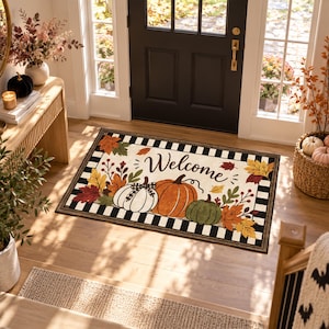 May include: A rectangular doormat featuring a black and white buffalo check border. The centre displays the word Welcome in black script, surrounded by illustrations of three pumpkins in white, orange, and green, accented with autumn leaves.