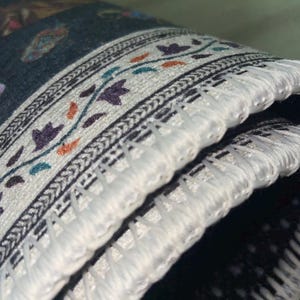 Pu&ograve; includere: Primo piano di un tappeto arrotolato con un bordo dettagliato. Il bordo presenta uno sfondo bianco con un motivo ripetuto di disegni floreali viola, arancioni e verde acqua. Il tappeto ha uno sfondo scuro e frange bianche.