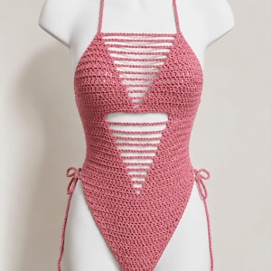 May include: A dusty rose crochet one-piece swimsuit featuring a halter neckline with thin straps, a triangular cutout design on the bodice with horizontal openwork detailing, and adjustable side ties at the hips for a customizable fit.