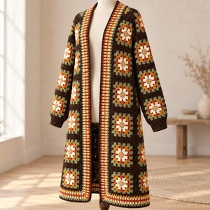Può includere: Un cardigan lungo e aperto realizzato con quadrati all'uncinetto marrone scuro. Ogni quadrato presenta un motivo floreale centrale nei toni del crema, arancione e verde salvia. Il capo ha maniche lunghe con polsini a coste e un bordo verticale.