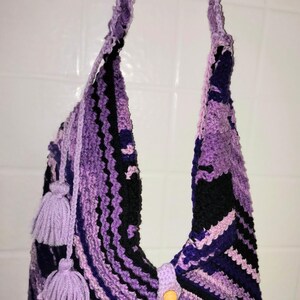 May include: A handmade crochet shoulder bag in shades of purple and black. The bag features a chevron pattern, a long strap, and two decorative tassels. A wooden button secures the flap.