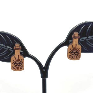 May include: A pair of wooden earrings featuring a bottle design with an eye symbol. The earrings are light brown with a dark brown eye and sunburst design. The earrings are displayed on a black metal stand with leaf-shaped accents.
