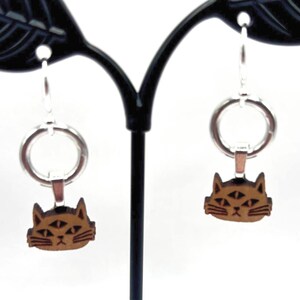 May include: A pair of earrings featuring a cat design. The earrings have a silver hoop and a brown cat face pendant. The cat has three eyes and is made of wood. The earrings are displayed on a black leaf-shaped earring stand.