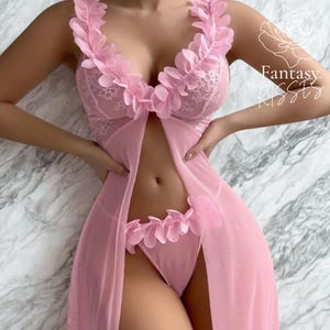 Könnte beinhalten: Rosa Dessous-Set mit einem transparenten Morgenmantel, einem BH mit Spitzenbesatz und passender Unterwäsche. Der Morgenmantel und die Unterwäsche sind mit gerüschten floralen Akzenten verziert. Der Text "Fantasy Kisses" ist rechts oben sichtbar.