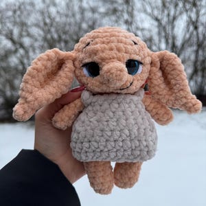 Puede incluir: Una muñeca de ganchillo hecha a mano con orejas grandes, ojos azules y un vestido gris. La muñeca es de color melocotón con un aspecto texturizado y afelpado. La muñeca se sostiene frente a un fondo borroso y nevado.