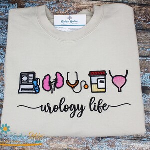 Puede incluir: Sudadera beige con bordados de temática urológica: ordenador, riñones, estetoscopio, frasco de pastillas y vejiga. Las palabras "urology life" están escritas en cursiva debajo.