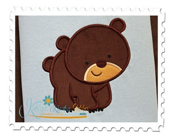 May include: Embroidered brown bear applique with a smiling face and a light brown nose. The bear is standing on a light blue fabric background.