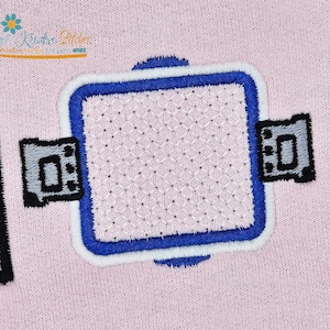 K&ouml;nnte beinhalten: Gestickter Aufn&auml;her mit quadratischem Design, blau-wei&szlig;em Rand und zwei grauen und schwarzen Seitendetails. Die Mitte des Quadrats hat ein Kreuzstichmuster, alles auf rosa Hintergrund.