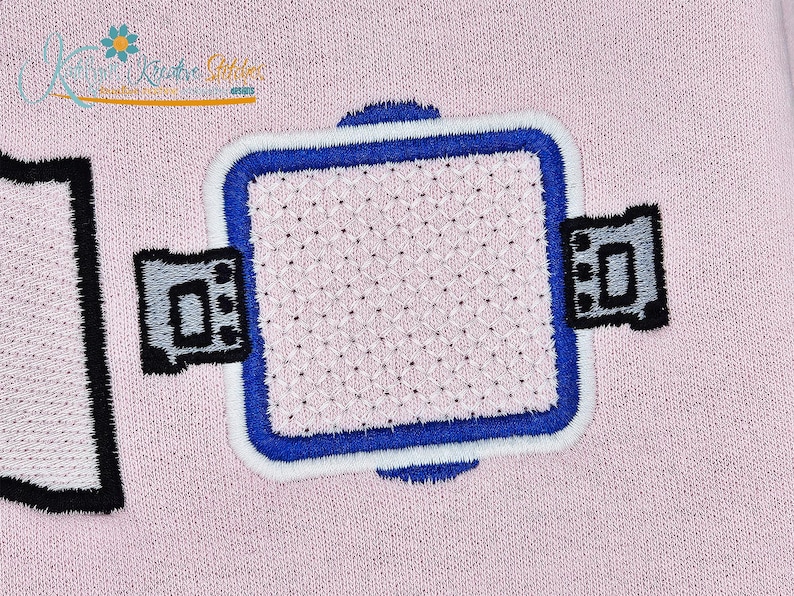 K&ouml;nnte beinhalten: Gestickter Aufn&auml;her mit quadratischem Design, blau-wei&szlig;em Rand und zwei grauen und schwarzen Seitendetails. Die Mitte des Quadrats hat ein Kreuzstichmuster, alles auf rosa Hintergrund.