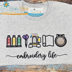 May include: A light gray t-shirt with an embroidered design featuring spools of thread, scissors, a sewing machine, fabric, and an embroidery hoop. The words "embroidery life" are written in cursive below the design.