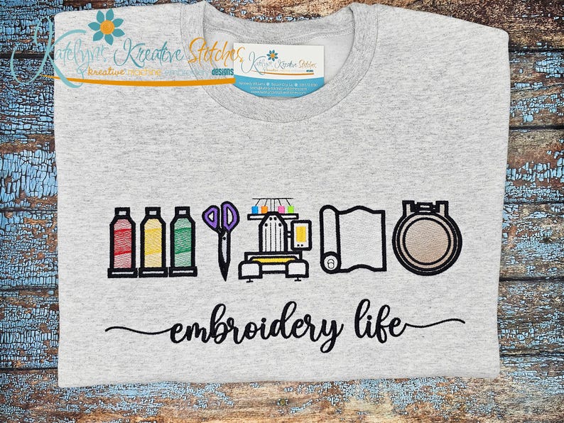 May include: A light gray t-shirt with an embroidered design featuring spools of thread, scissors, a sewing machine, fabric, and an embroidery hoop. The words "embroidery life" are written in cursive below the design.
