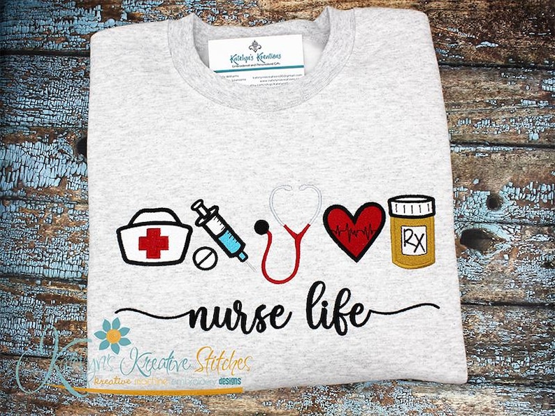 May include: A gray t-shirt with an embroidered design of a nurse's cap, syringe, stethoscope, heart with a heartbeat line, and a pill bottle with "RX" on it. The text "nurse life" is embroidered below the design.