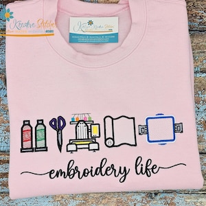 K&ouml;nnte beinhalten: Hellrosa Sweatshirt mit dem Schriftzug "embroidery life" in Schreibschrift. Das Design umfasst gestickte Bilder von N&auml;hwerkzeugen, darunter Flaschen, Schere, N&auml;hmaschine, Stoff und einen Stickrahmen.