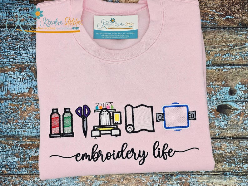 K&ouml;nnte beinhalten: Hellrosa Sweatshirt mit dem Schriftzug "embroidery life" in Schreibschrift. Das Design umfasst gestickte Bilder von N&auml;hwerkzeugen, darunter Flaschen, Schere, N&auml;hmaschine, Stoff und einen Stickrahmen.
