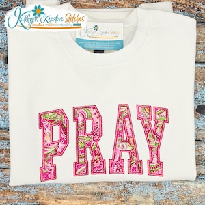 Op de afbeelding: Een wit sweatshirt met ronde hals en het woord PRAY in grote applicatieletters. De letters zijn gevuld met een roze, groen en wit paisley-patroon en omlijnd met een dikke roze stikselrand. Een merklabel bevindt zich bij de halslijn.