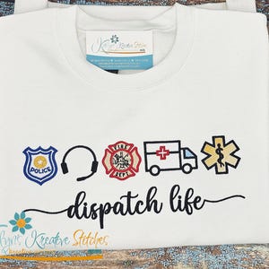 Puede incluir: Sudadera blanca con símbolos bordados de policía, auriculares, bomberos, ambulancia y estrella médica. Las palabras "dispatch life" están escritas en guión negro debajo de los símbolos.