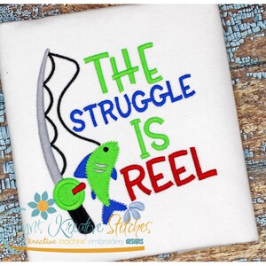 Może przedstawiać: Haftowany wzór przedstawiający wędkę i rybę z tekstem "The Struggle Is Reel" w kolorze zielonym, niebieskim i czerwonym.