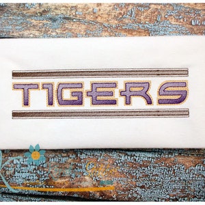 May include: White fabric with the word "TIGERS" embroidered in purple and gold letters with brown outlines. The letters are outlined with two brown lines.