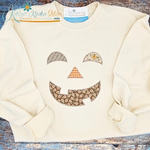 May include: A cream-colored long-sleeve ribbed sweatshirt featuring a jack-o'-lantern face applique. The eyes, nose, and mouth are crafted from various brown, orange, and floral patterned fabrics with visible stitching around the edges.