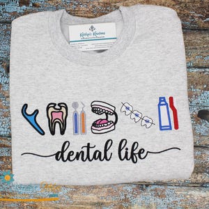 Puede incluir: Sudadera gris claro con gráficos bordados con temática dental. El diseño incluye un diente, instrumentos dentales, aparatos ortopédicos y un cepillo de dientes, con las palabras "dental life" en una fuente de escritura. Una etiqueta es visible en el cuello.