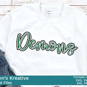 Op de afbeelding: Wit sweatshirt met het woord "Demons" in groene en zwarte letters. De tekst is in een cursief lettertype. De afbeelding is op een houten achtergrond.