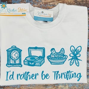 May include: White sweatshirt featuring turquoise embroidered images of a clock, record player, hen, and decorative spoons. The text "I'd rather be thrifting" is embroidered below the images.