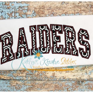 Könnte beinhalten: Weißer Stoff mit dem Wort "RAIDERS", das in schwarz-weißen geometrischen Mustern mit roten Akzenten gestickt ist. Der Text ist in einer fetten, gebogenen Schriftart gehalten. Das Bild befindet sich auf einer verwitterten, blau gestrichenen Holzoberfläche.