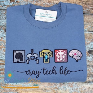 Op de afbeelding: Een blauwe crewneck sweatshirt met geborduurde medische afbeeldingen. Het ontwerp bevat een schedel, skelet en hersenen, met de tekst "xray tech life" in een scriptlettertype. Een label is bevestigd bij de halslijn.