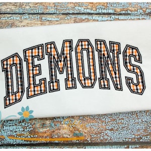 May include: White t-shirt featuring the word "DEMONS" in an arched design. The letters are filled with a black, orange, and white plaid pattern, outlined with black stitching. The shirt is presented on a distressed, blue and brown wooden surface.