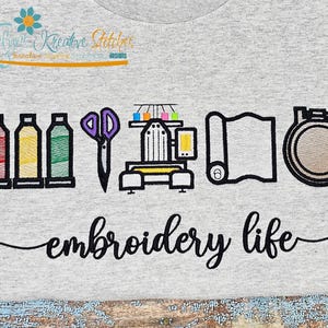 May include: A light gray t-shirt featuring an embroidery design with spools of thread, scissors, an embroidery machine, fabric, and a hoop. The words "embroidery life" are written in cursive below the design.