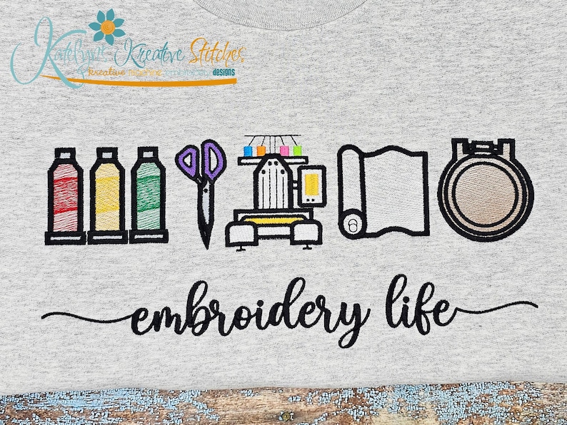 May include: A light gray t-shirt featuring an embroidery design with spools of thread, scissors, an embroidery machine, fabric, and a hoop. The words "embroidery life" are written in cursive below the design.
