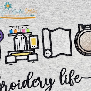 May include: Embroidered design on a light gray fabric. The design includes scissors, an embroidery machine with colorful thread spools, a roll of fabric, and an embroidery hoop. The words "embroidery life" are written in a script font.