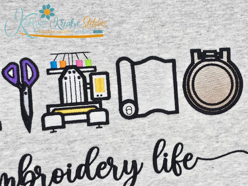 May include: Embroidered design on a light gray fabric. The design includes scissors, an embroidery machine with colorful thread spools, a roll of fabric, and an embroidery hoop. The words "embroidery life" are written in a script font.