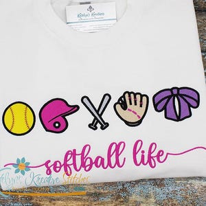 May include: White t-shirt with a pink, purple and yellow softball design. The design includes a softball, a helmet, bats, a glove and a bow. The text "softball life" is written in pink below the design.