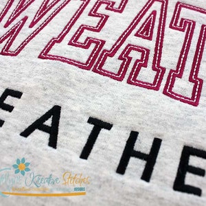 May include: Close-up of a gray sweatshirt with the word "SWEATER" embroidered in pink thread and the word "WEATHER" embroidered in black thread.