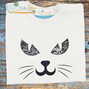 Könnte beinhalten: Ein weißes Sweatshirt mit Rundhalsausschnitt und einem gestickten Katzengesicht. Die Augen bestehen aus schwarzem Spitzenstoff mit komplizierten Mustern, während die Nase und die Schnurrhaare mit schwarzem Garn und Paisley-Muster gestickt sind.