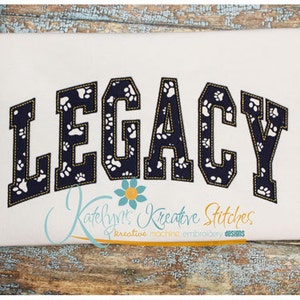 May include: A white fabric with the word "LEGACY" in navy blue letters with a white paw print pattern. The letters have a yellow outline and are in a varsity style font.