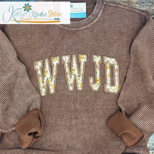 May include: A brown long-sleeve waffle-knit sweatshirt featuring the letters WWJD appliqued in a cream fabric with a yellow and green floral pattern. The letters are outlined in a brown thread, and the garment has a ribbed crew neckline.