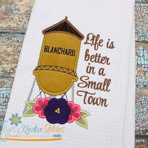 May include: Embroidered design featuring a water tower with the text "BLANCHARD" and the phrase "Life is better in a small town". The design includes a floral motif with pink, purple, and green flowers.