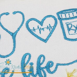 May include: Embroidered design featuring a stethoscope, a heart with a heartbeat line, and a pill bottle with an RX symbol. The text "a life" is also embroidered in blue thread.