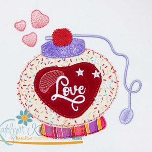May include: An embroidered design featuring a snow globe with a red heart that says "Love". The snow globe has a purple lid and is decorated with sprinkles and stars. Three small hearts float above the globe.