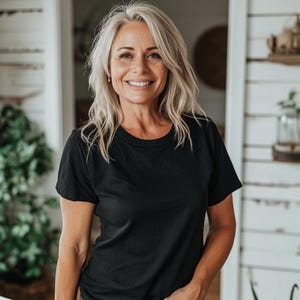 May include: A woman with gray hair smiles while wearing a black crew neck t-shirt. The t-shirt is a solid black color and appears to be made of a soft material. The background includes a white door and a glimpse of a shelf with decorative items.