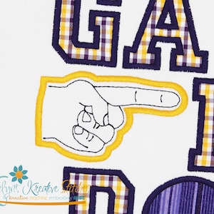 May include: White fabric with embroidered letters and a pointing hand. The letters are in a purple and yellow plaid pattern with a dark blue outline. The pointing hand is outlined in yellow.