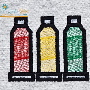 May include: Embroidered design featuring three colorful paint tubes in red, yellow, and green, outlined in black. The design is on a light gray fabric, with the text "Kreative Stitches" in the upper left corner.
