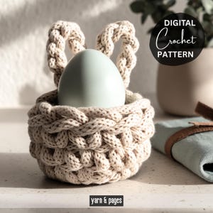 May include: A beige crocheted basket with two looped handles, holding a pale blue egg. The basket has a textured, woven appearance. The words "DIGITAL Crochet PATTERN" are in a black circle. The text "yarn & pages" is at the bottom.