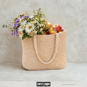 Könnte beinhalten: Eine beige gehäkelte Tragetasche gefüllt mit einem Strauß weißer Gänseblümchen, lila Blumen und anderem Grünzeug, zusammen mit mehreren roten und gelben Äpfeln. Die Tasche hat zwei Griffe und steht auf einer hellen Oberfläche.