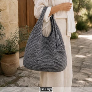 Peut inclure: Un sac hobo en crochet gris ardoise avec un motif de mailles texturé, une large bandoulière intégrée et un long pompon assorti suspendu sur le côté. Le bas de l'image affiche l'étiquette de texte yarn & pages dans un encadré noir.