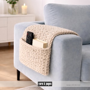 Peut inclure: Un organiseur de bras de canapé en crochet de couleur crème avec une poche. Une télécommande noire et une petite boîte beige se trouvent à l'intérieur de la poche. L'organisateur est drapé sur le bras d'un canapé bleu clair. Le texte "yarn & pages" est en bas.